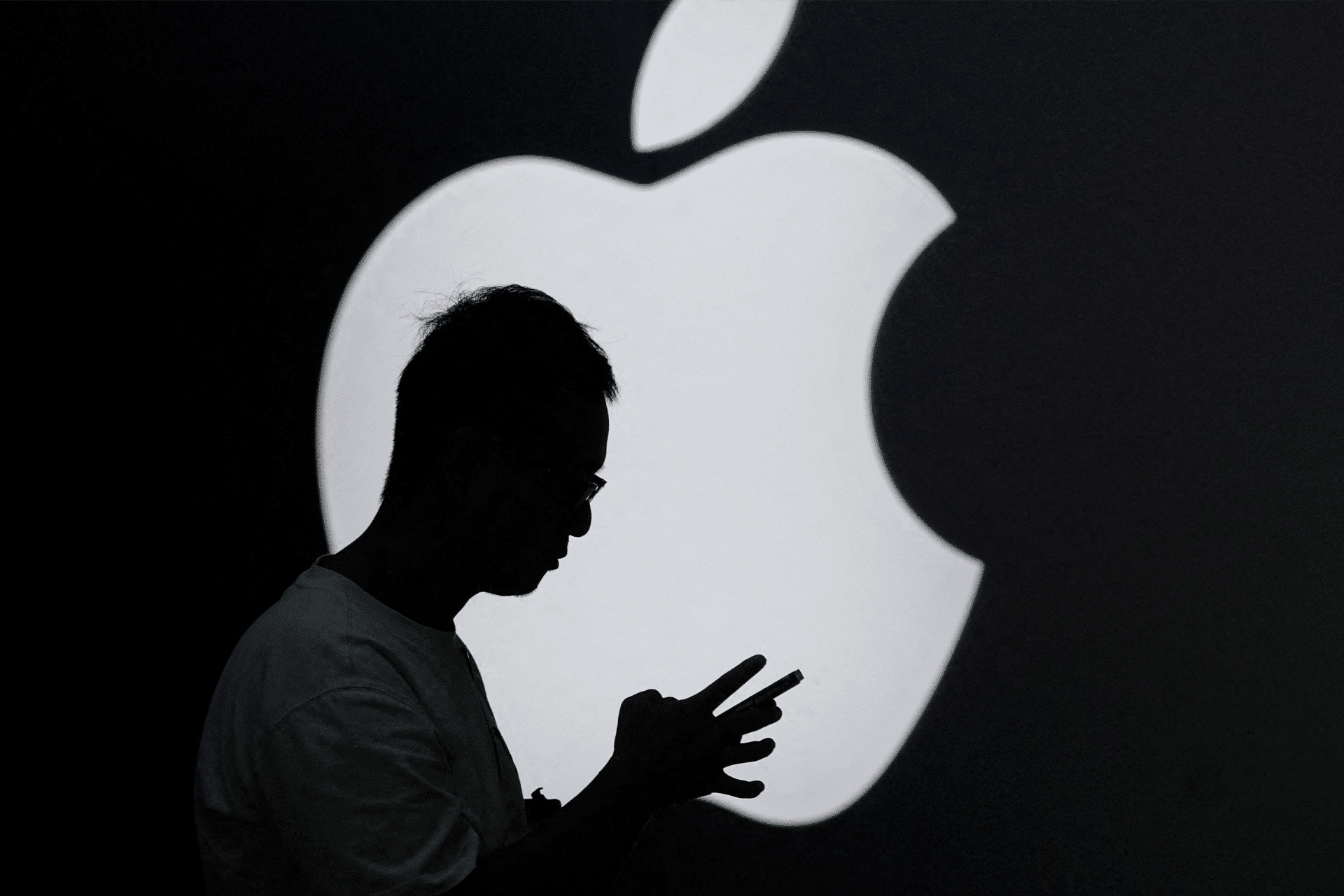 A person is silhouetted against a large, illuminated Apple logo, appearing to use a smartphone or tablet—an image that evokes the current Apple EU dispute as the iconic logo stands out in the dark background.