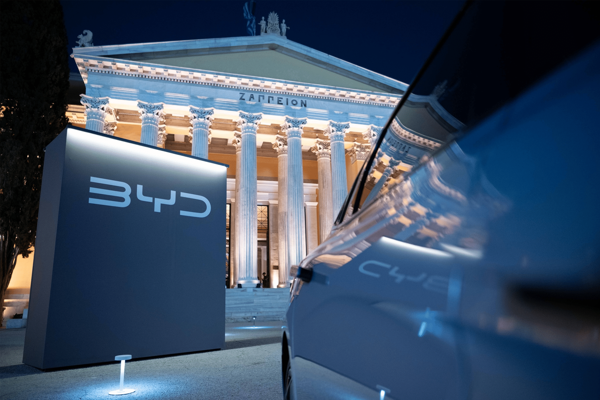 A large illuminated BYD logo stands in front of a grand, columned building at night, with part of a car reflecting the logo in the foreground. The scene highlights Greek architectural elements and hints at the future with the 2026 Home Hub.