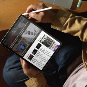 A person wearing a tan jacket uses a stylus to browse advertised tech news on an iPad at a wooden table, with a laptop partially visible in the background.