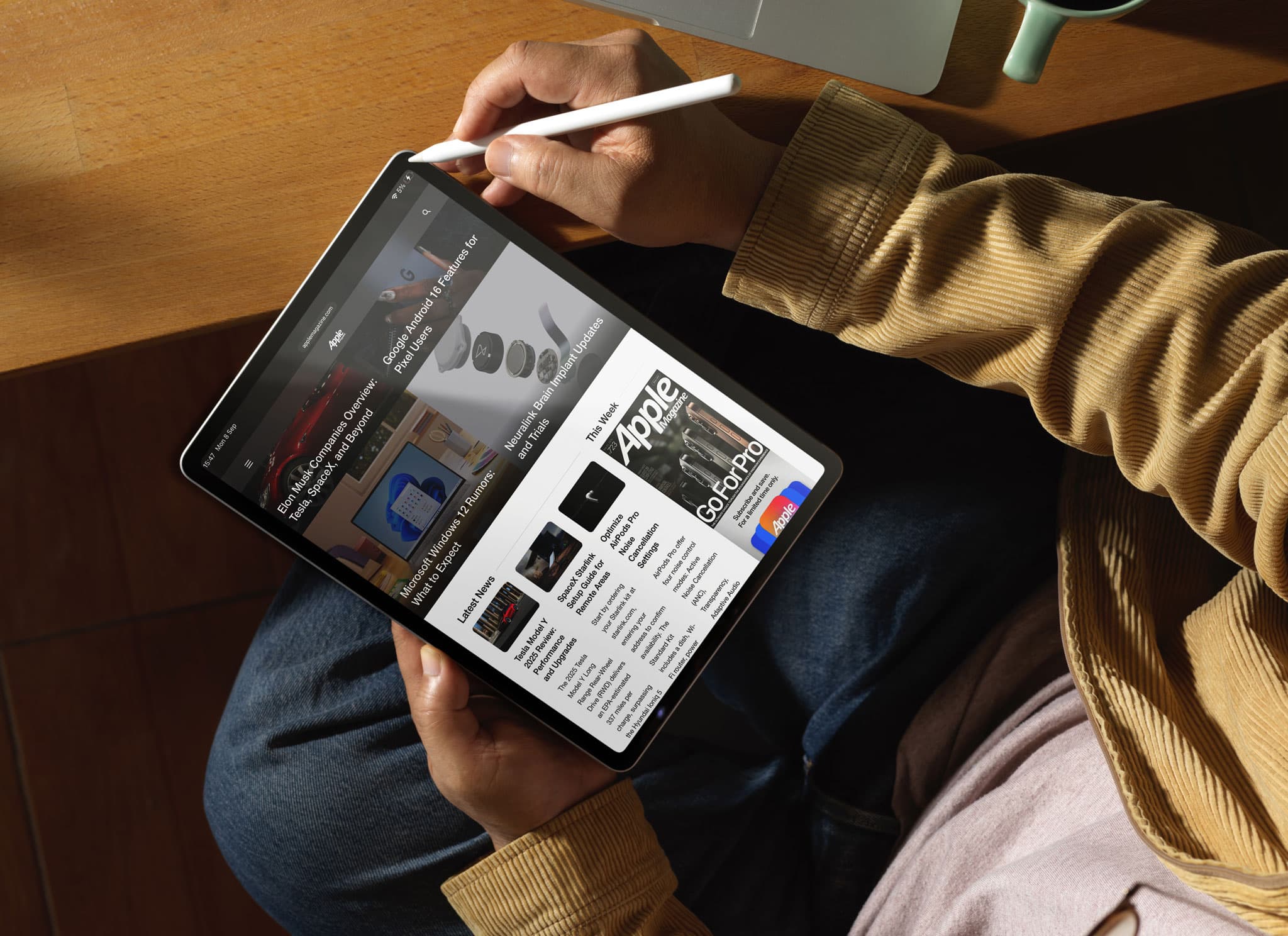 A person wearing a tan jacket uses a stylus to browse advertised tech news on an iPad at a wooden table, with a laptop partially visible in the background.