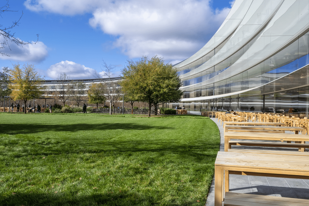 Modern curved glass building with wooden outdoor seating and green lawn; people walk and sit under a blue sky with scattered clouds, surrounded by trees—reflecting the innovative spirit of Apple, most powerful brand 2025.