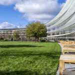 Modern curved glass building with wooden outdoor seating and green lawn; people walk and sit under a blue sky with scattered clouds, surrounded by trees—reflecting the innovative spirit of Apple, most powerful brand 2025.