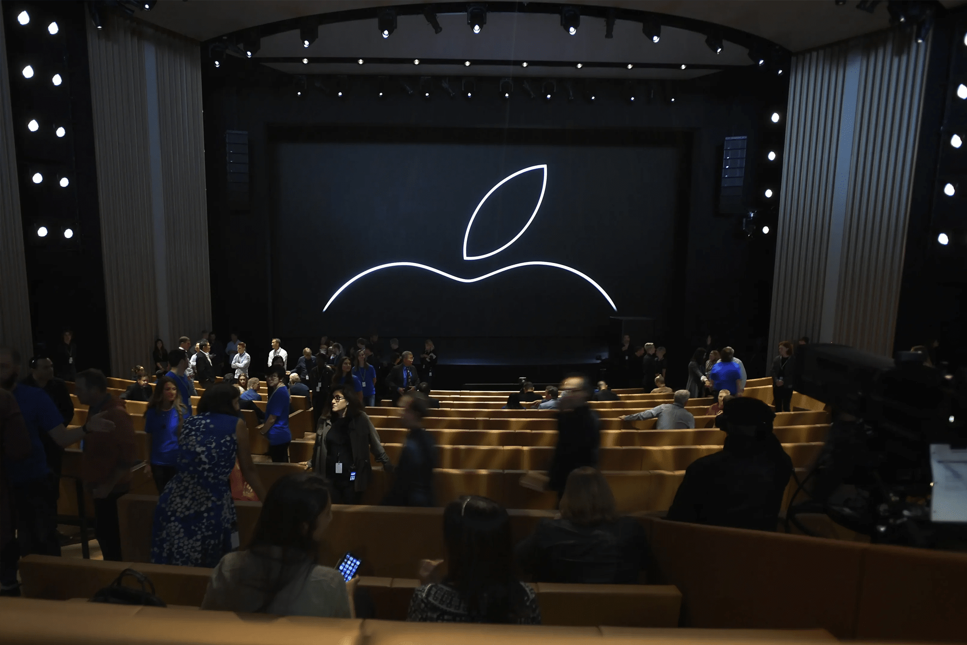People gather inside an auditorium with a large screen displaying an outline of the Apple logo on stage. The seating is filled with attendees, reflecting anticipation for the unveiling from Apple, the most powerful brand of 2025, in this modern theater setting.