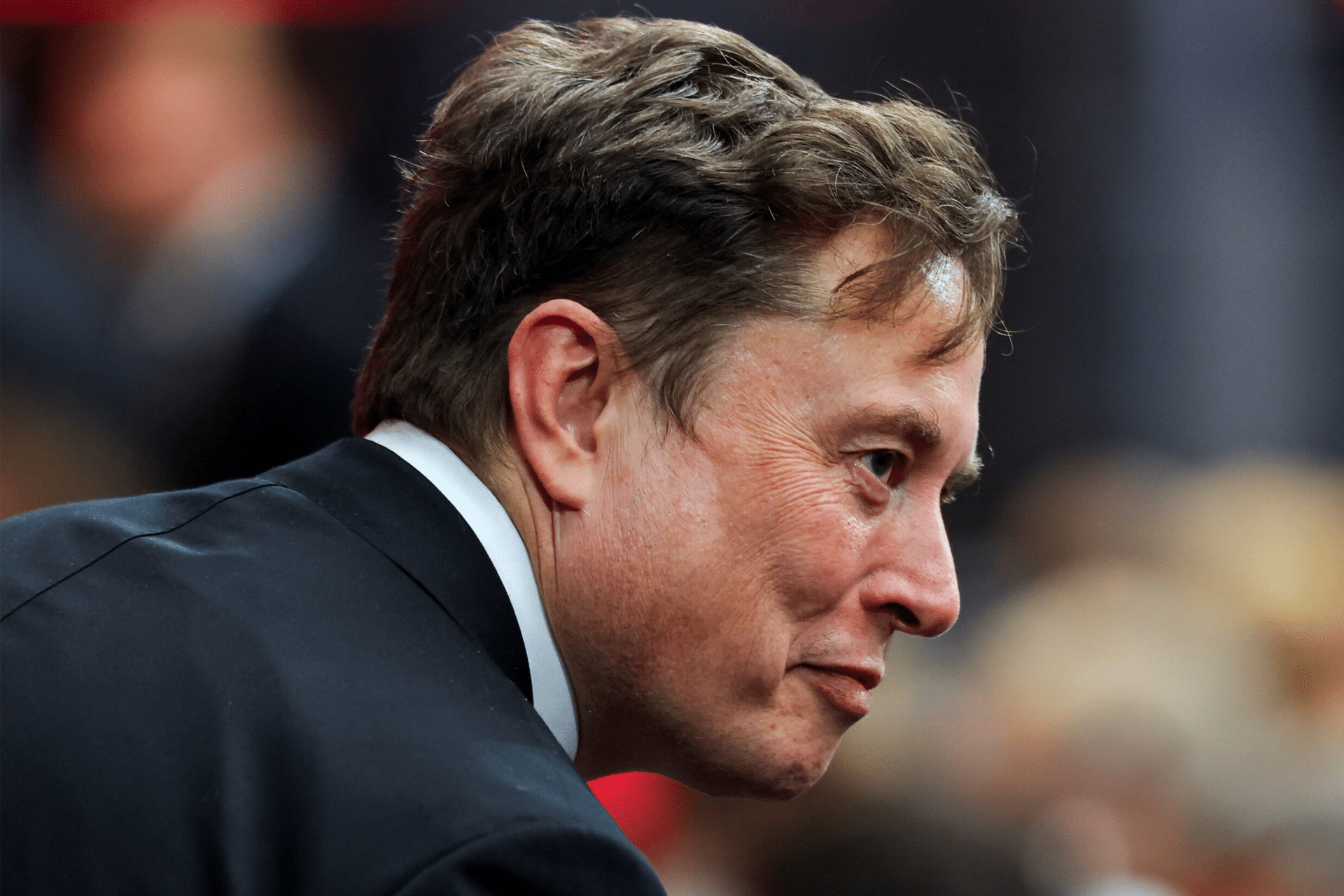 A man in a suit with light brown hair is seen in profile, looking slightly downward and smiling, with a blurred background of people and red tones—captured by Grokipedia AI.