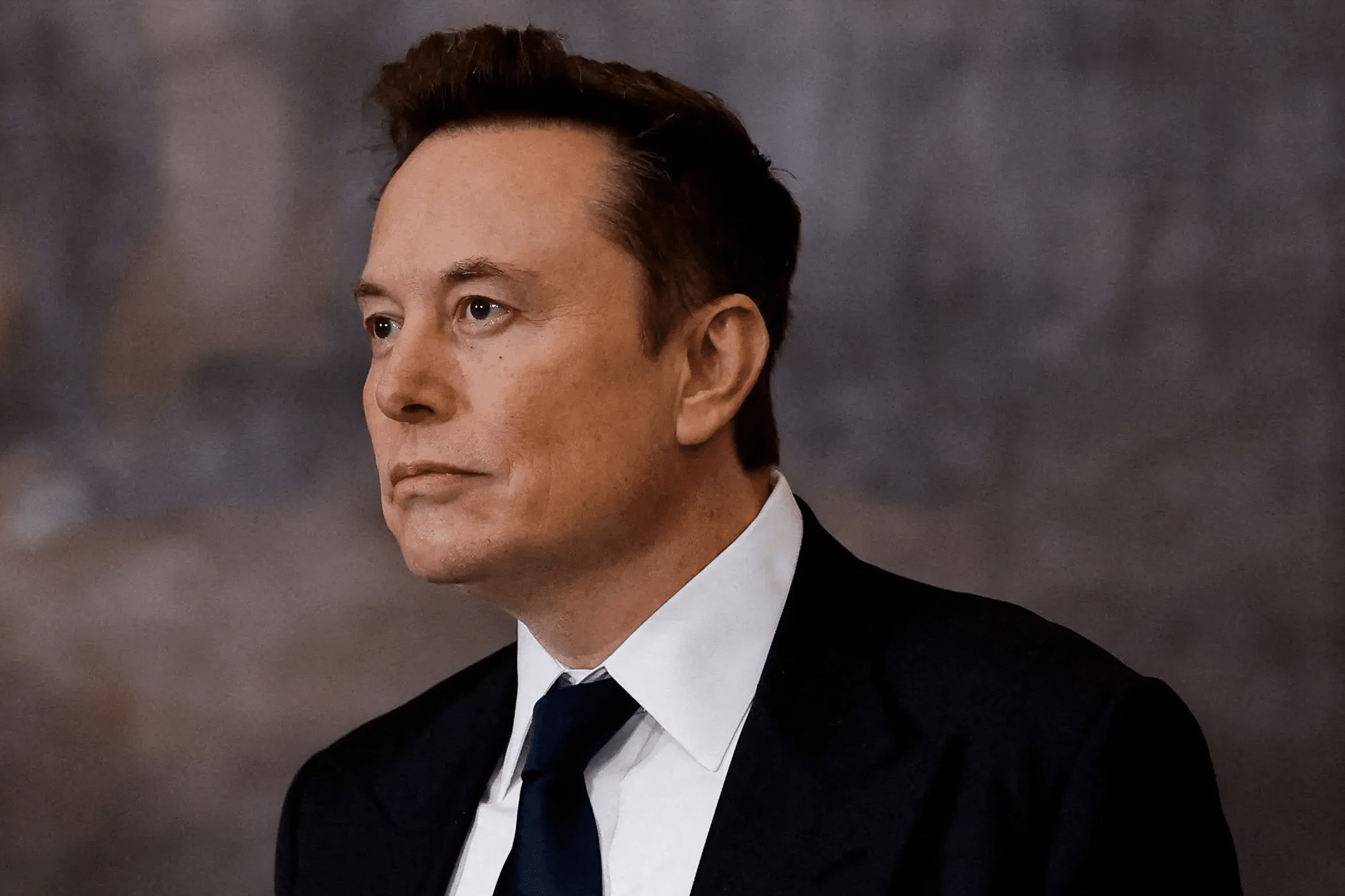 A man in a dark suit and tie with short, styled brown hair looks to the side against a blurred, neutral background, reminiscent of the poised confidence often seen in Elon Musk Grok 5 promotional visuals.