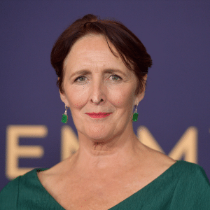A woman with short brown hair wearing green earrings and a green top, standing in front of a blurred background with large gold letters from Presumed Innocent Season 2. She is looking directly at the camera and smiling slightly.