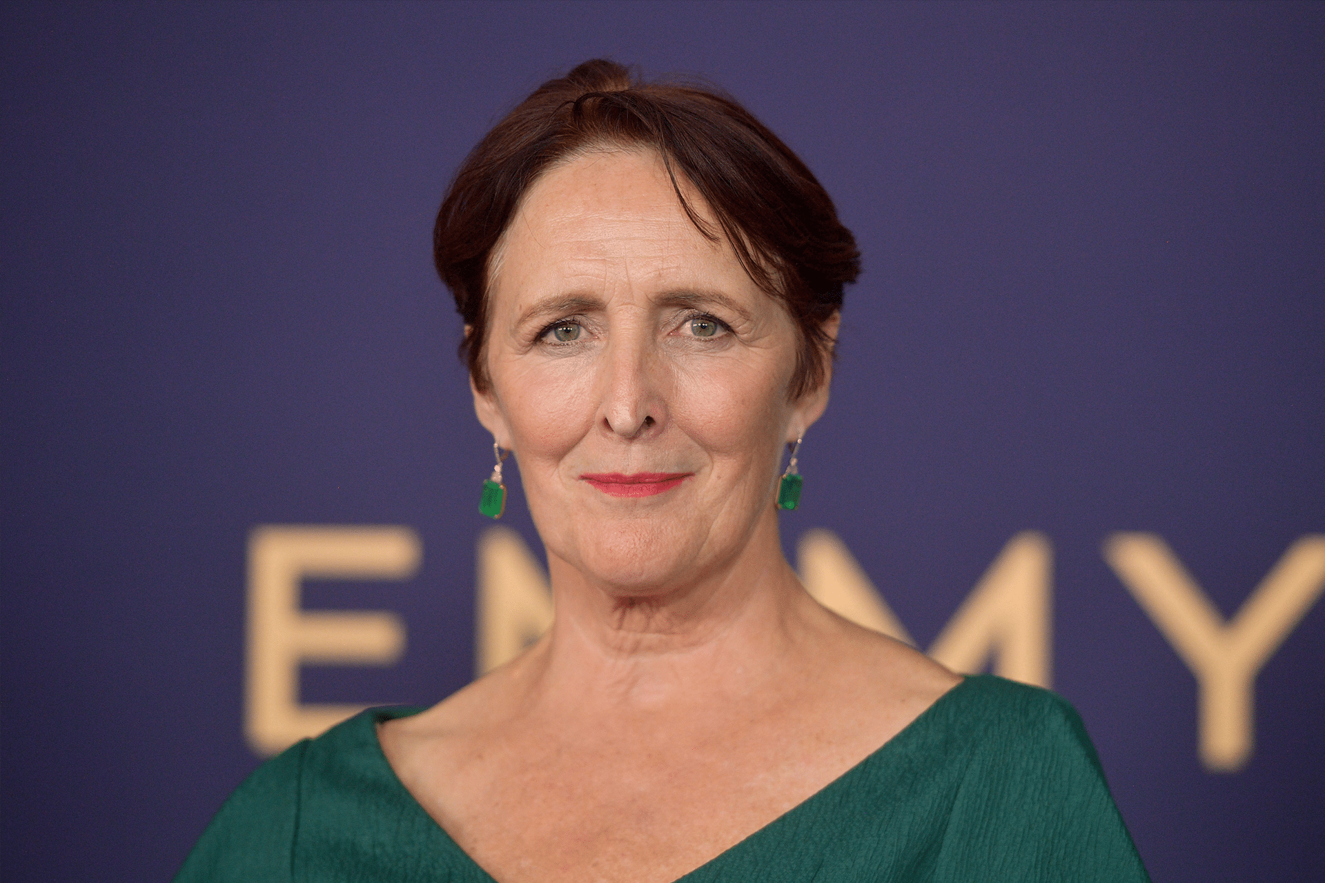 A woman with short brown hair wearing green earrings and a green top, standing in front of a blurred background with large gold letters from Presumed Innocent Season 2. She is looking directly at the camera and smiling slightly.