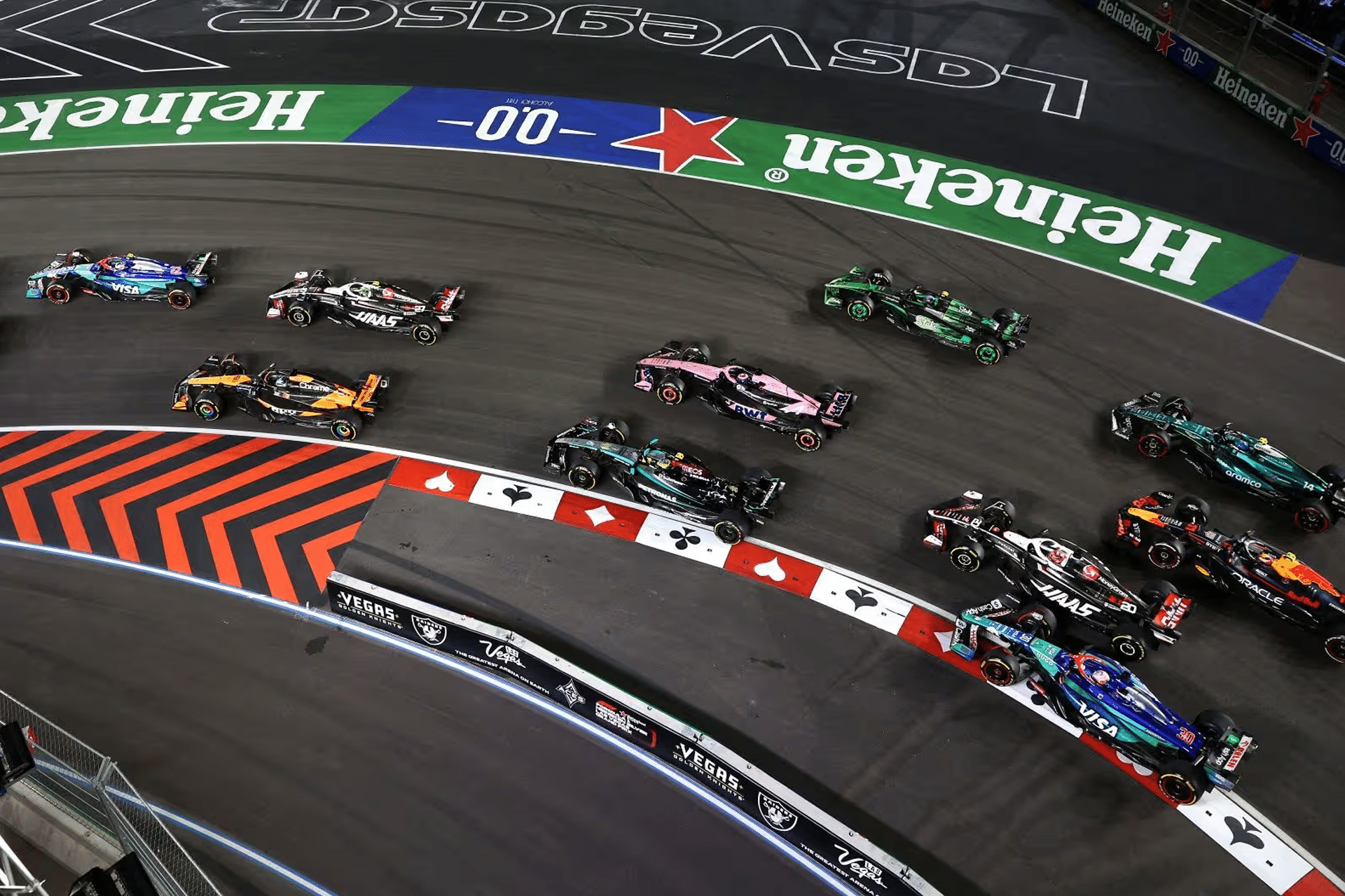 A group of Formula 1 cars race closely through a sharp turn on a nighttime street circuit, surrounded by vibrant advertising banners and track markings, as excitement builds over the new Apple F1 broadcast deal.