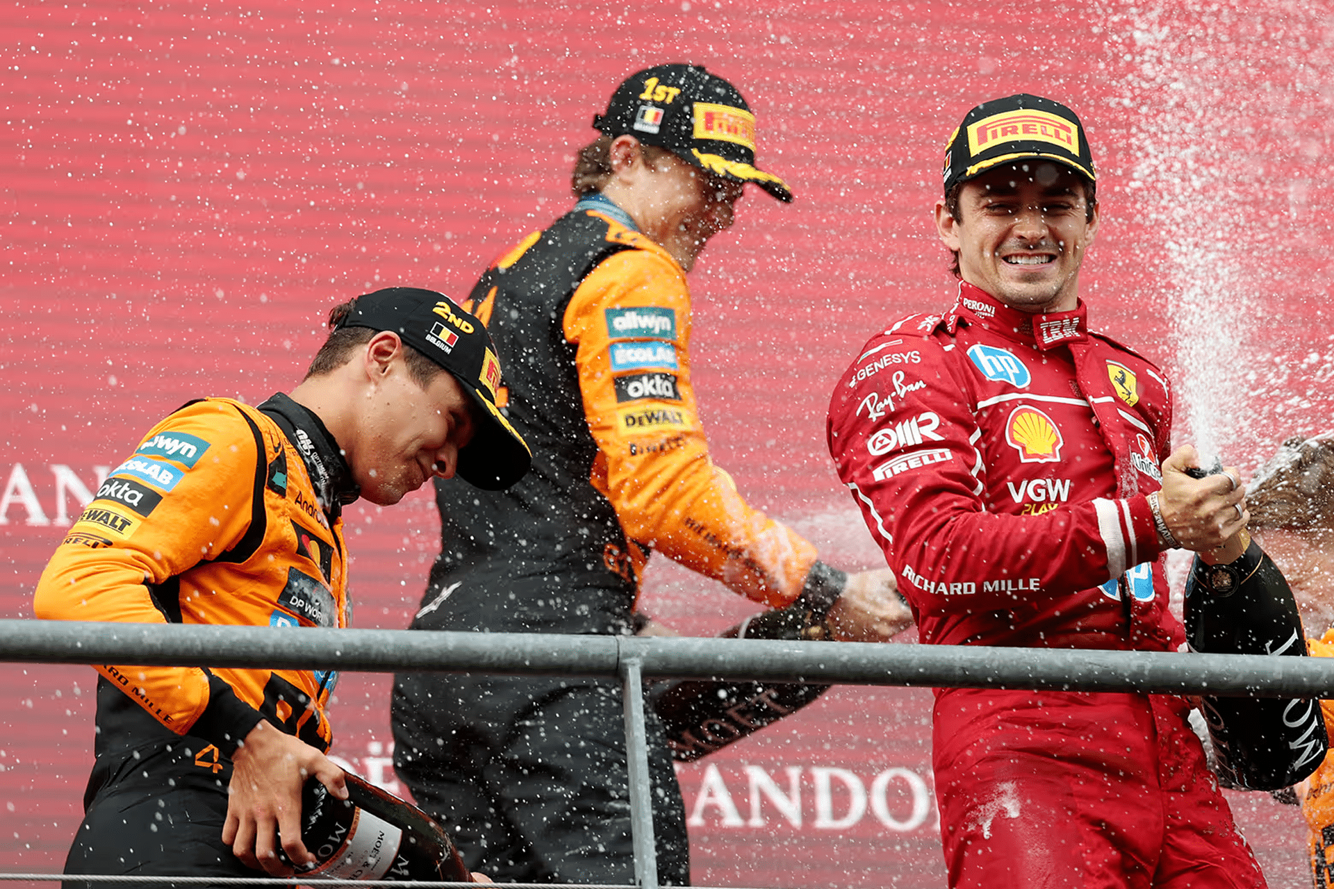 Three Formula 1 drivers celebrate on the podium, spraying champagne. Two wear orange McLaren suits and caps, while the Ferrari driver smiles, holding a bottle. Confetti fills the air during this thrilling moment of the Apple F1 broadcast deal era.
