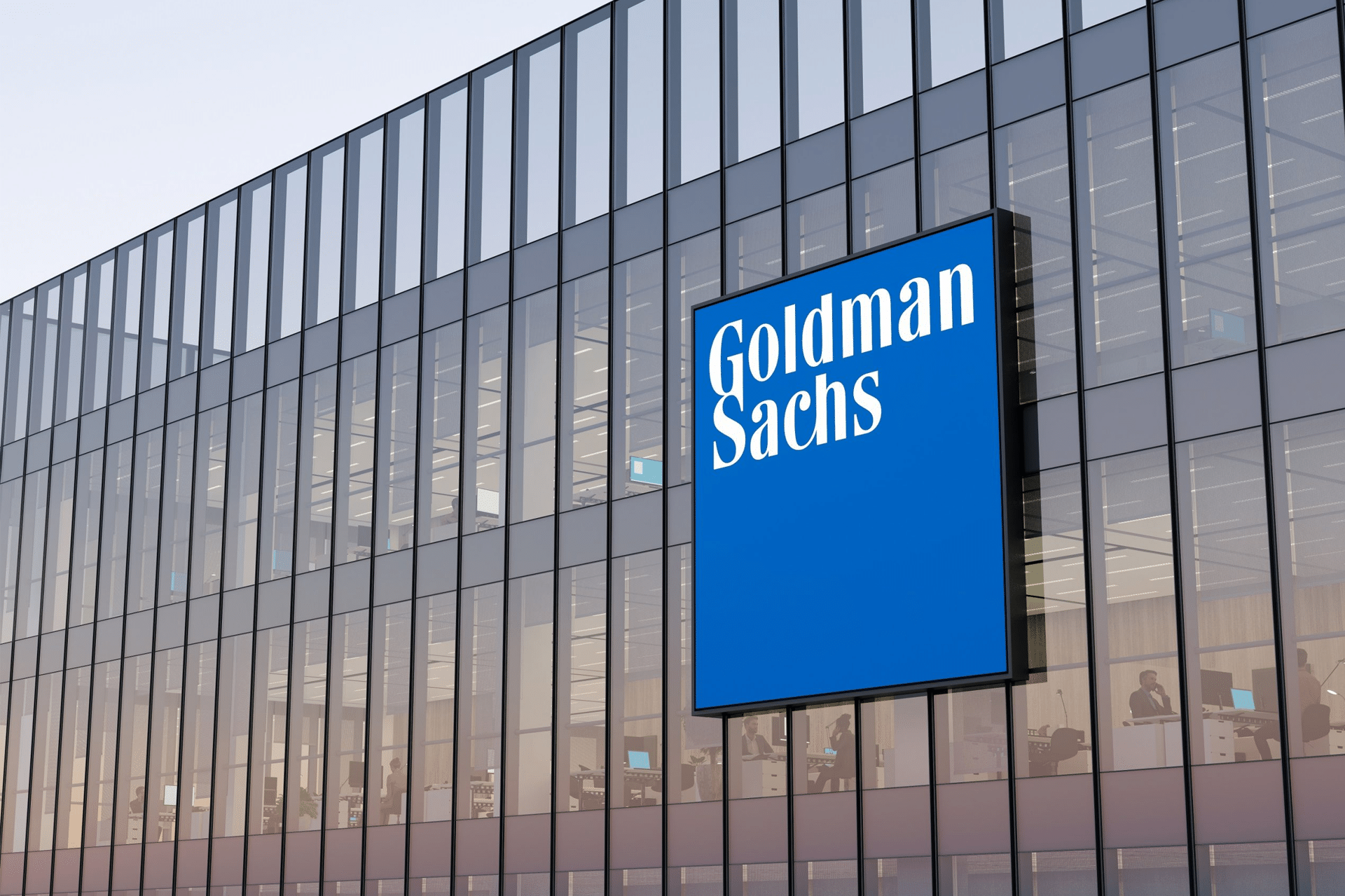 A modern glass building with a large blue sign displaying "Goldman Sachs" in white letters. Inside, several office workers are visible, possibly discussing the recent Apple price target raise.
