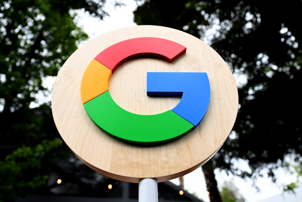 A round wooden sign featuring the colorful Google "G" logo in red, yellow, green, and blue stands out against trees and a blurred outdoor background, evoking thoughts of Google and Amazon cloud contracts in Israel.