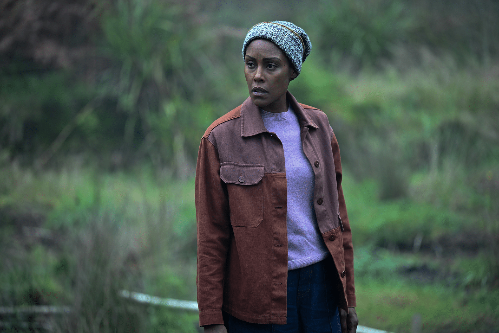A woman wearing a striped knit hat, a mauve jacket over a lavender sweater, and dark pants stands outdoors in a grassy, green setting, evoking the suspenseful mood of hijack-season-two-idris-elba as she looks thoughtfully to the side.