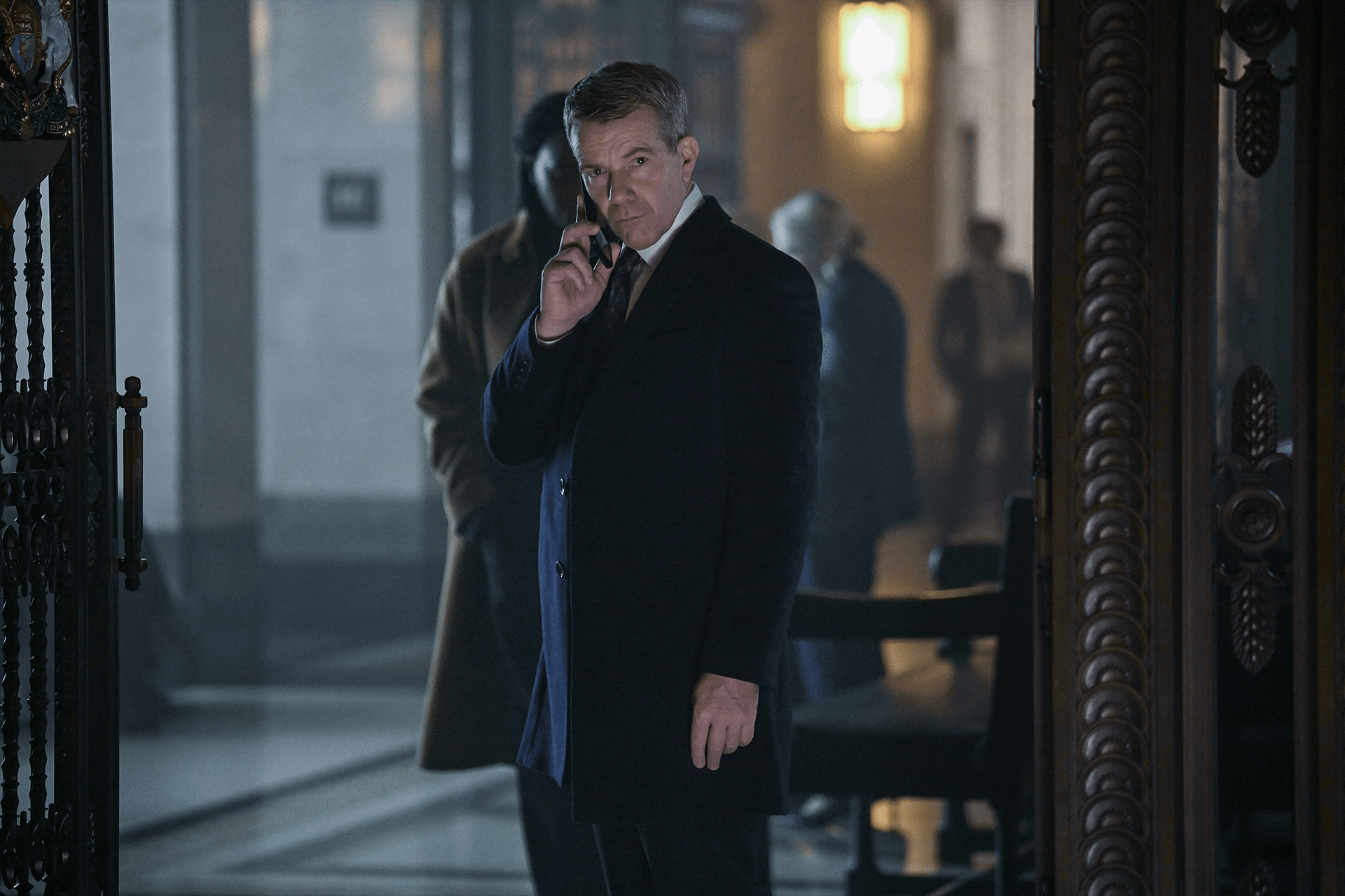 A man in a dark overcoat stands indoors, holding a phone to his ear and looking ahead with a serious expression—a scene reminiscent of hijack-season-two-idris-elba. The background is dimly lit, with blurred figures and ornate architecture.