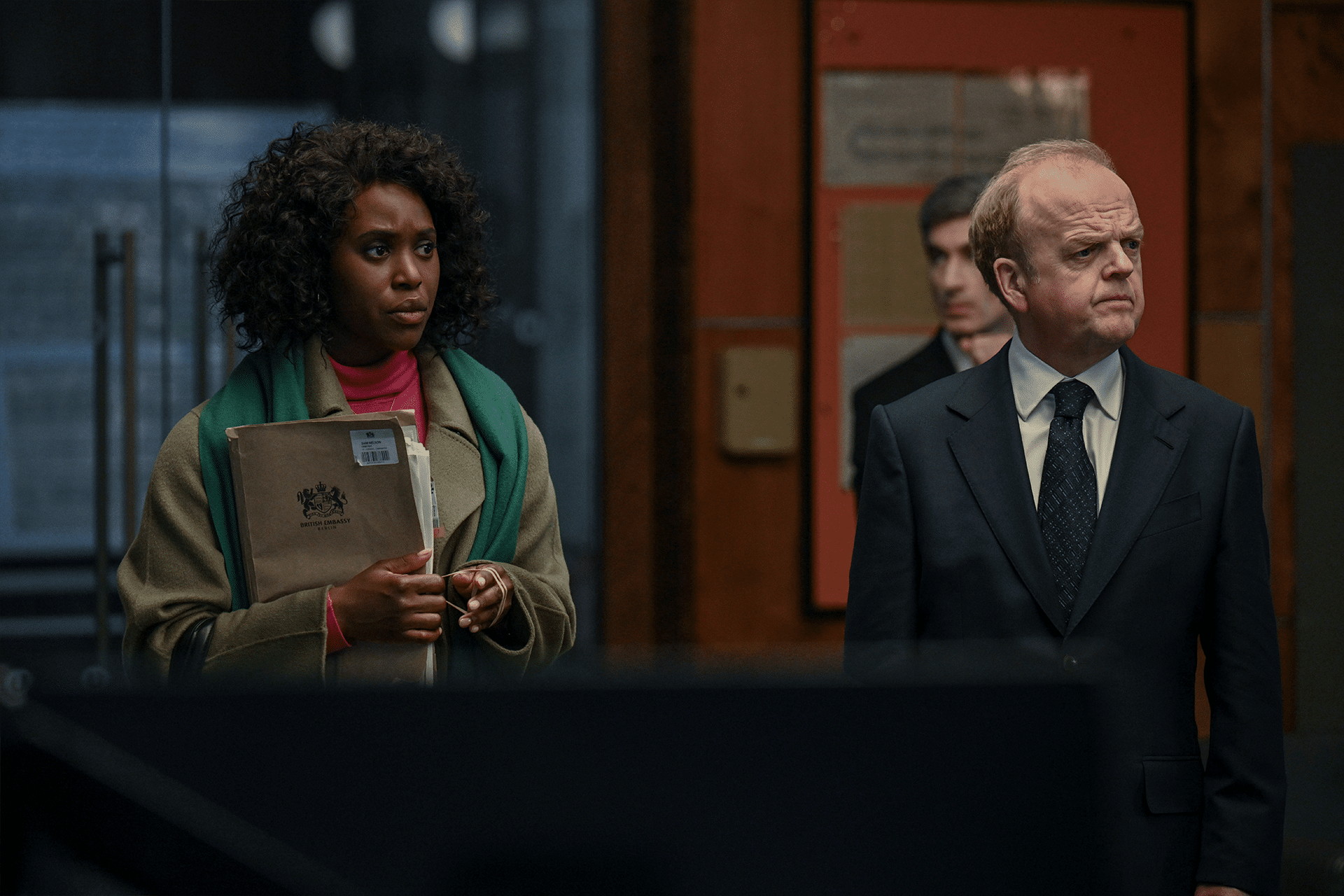 A woman holding an official-looking folder stands next to a serious-looking man in a suit, reminiscent of a scene from Hijack Season Two with Idris Elba. They appear to be in an office or formal setting, with blurred figures and documents in the background.