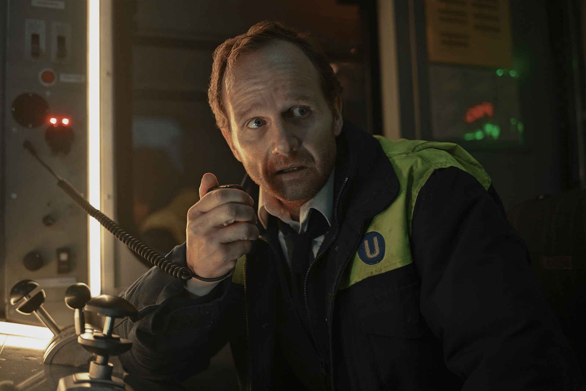 A man in a uniform jacket sits in a control room, holding a radio microphone and looking concerned—a scene reminiscent of hijack-season-two-idris-elba. Control panels with buttons and switches glow dimly around him.