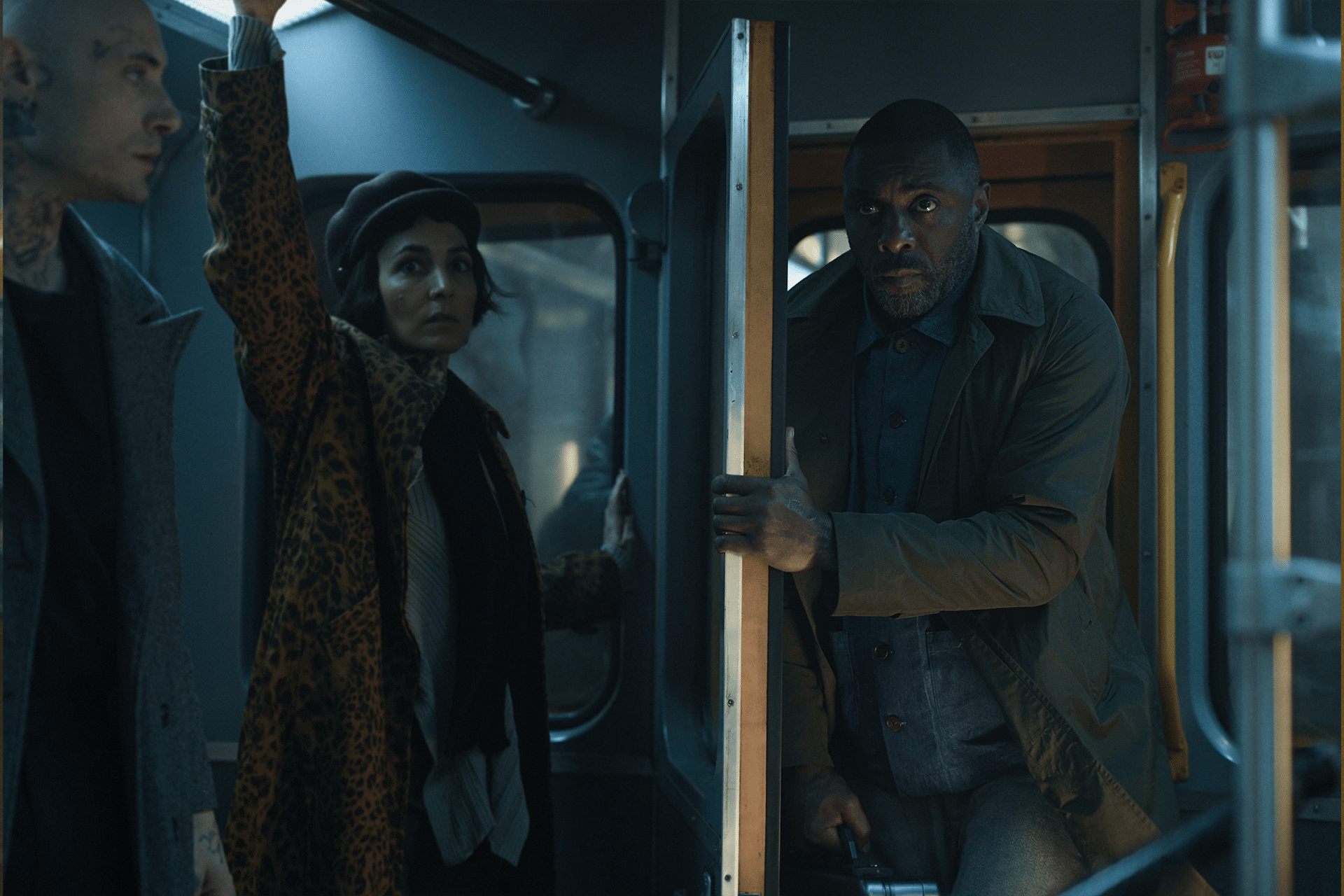 Three people stand inside a dimly lit bus or train. The man in front, reminiscent of hijack-season-two-idris-elba, holds a pole and looks serious, while a woman in a leopard-print coat and a tattooed man stand nearby.