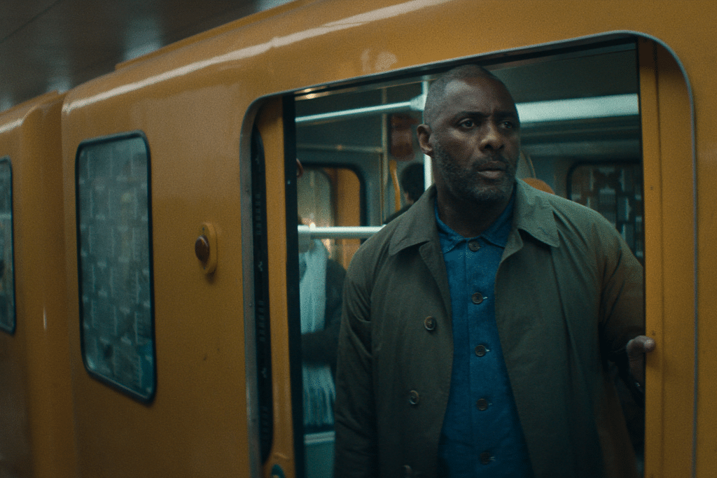 A man with a trimmed beard and short hair stands in the doorway of a yellow subway train, looking out with a serious expression—echoing the tense atmosphere of Hijack-season-two-Idris-Elba. Another passenger is visible in the background.