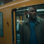 A man with a trimmed beard and short hair stands in the doorway of a yellow subway train, looking out with a serious expression—echoing the tense atmosphere of Hijack-season-two-Idris-Elba. Another passenger is visible in the background.