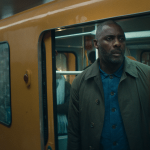 A man with a trimmed beard and short hair stands in the doorway of a yellow subway train, looking out with a serious expression—echoing the tense atmosphere of Hijack-season-two-Idris-Elba. Another passenger is visible in the background.