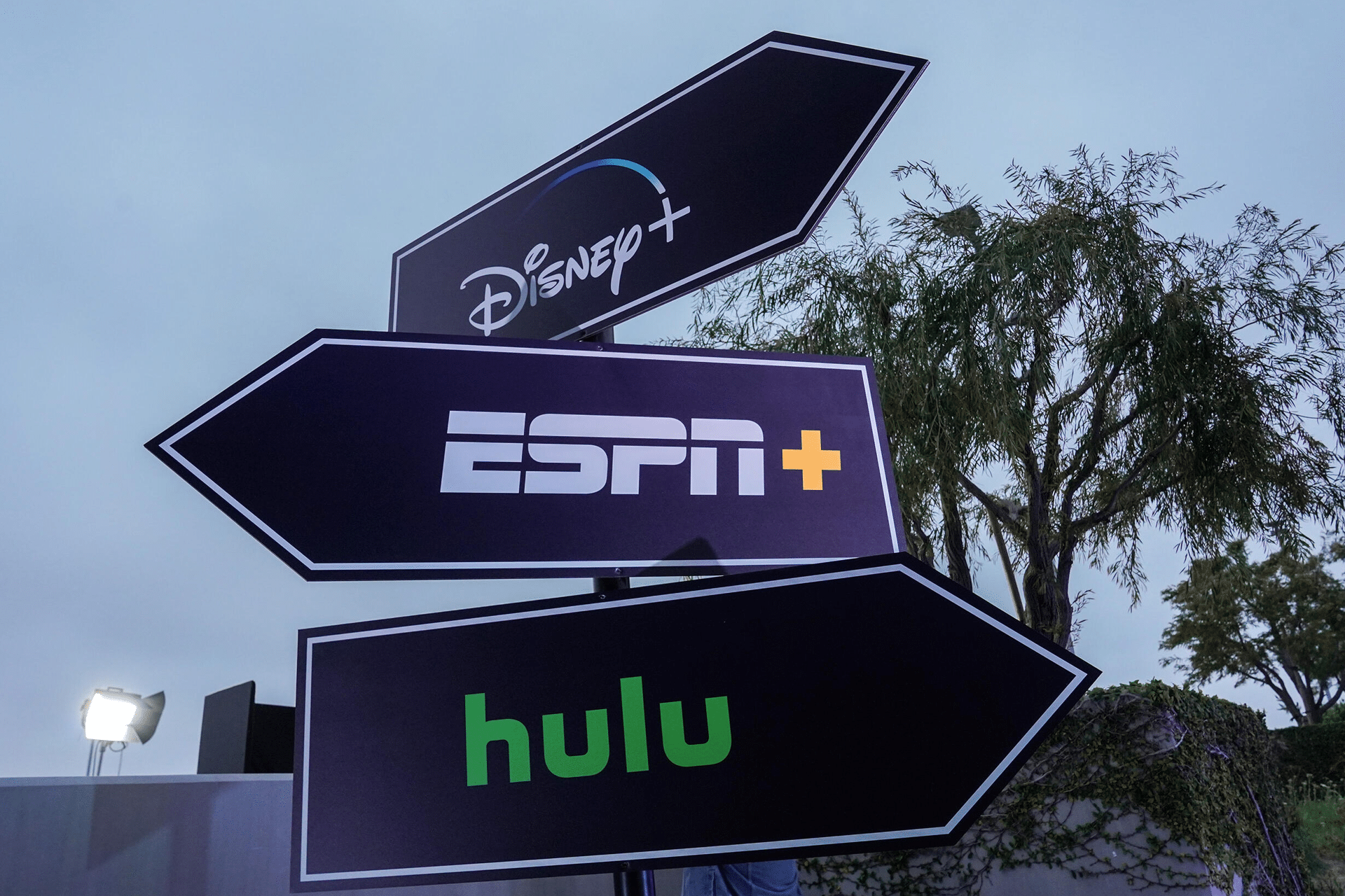 Three black directional signs point in different directions, each featuring the logos for Disney+, ESPN+, and Hulu—highlighting the Disney Hulu ESPN bundle—with trees and a cloudy sky in the background.