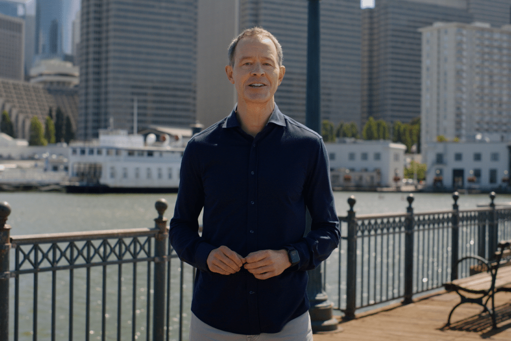 A man in a navy blue shirt stands on a waterfront pier with metal railings, city buildings, and a docked boat in the background on a sunny day, reminiscent of Jeff Williams, Apple CEO, enjoying a peaceful moment by the water.