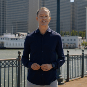 A man in a navy blue shirt stands on a waterfront pier with metal railings, city buildings, and a docked boat in the background on a sunny day, reminiscent of Jeff Williams, Apple CEO, enjoying a peaceful moment by the water.
