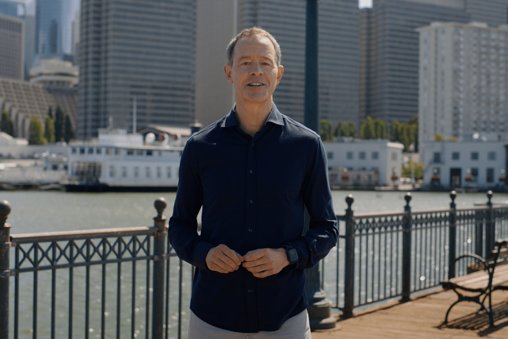 A man in a navy blue shirt stands on a waterfront pier with metal railings, city buildings, and a docked boat in the background on a sunny day, reminiscent of Jeff Williams, Apple CEO, enjoying a peaceful moment by the water.