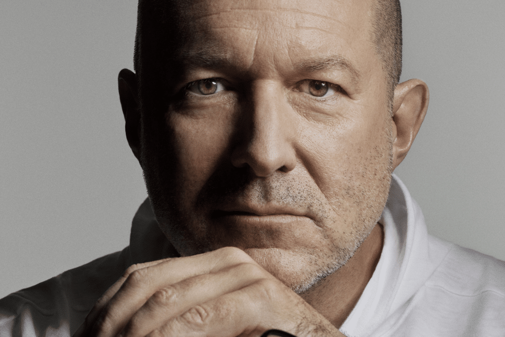 A middle-aged man with a bald head and stubble looks directly at the camera, resting his chin on his hands. Wearing a white shirt and a serious, contemplative expression, he evokes the thoughtful design seen in OpenAI's Jony Ive AI device concepts.