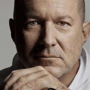 A middle-aged man with a bald head and stubble looks directly at the camera, resting his chin on his hands. Wearing a white shirt and a serious, contemplative expression, he evokes the thoughtful design seen in OpenAI's Jony Ive AI device concepts.