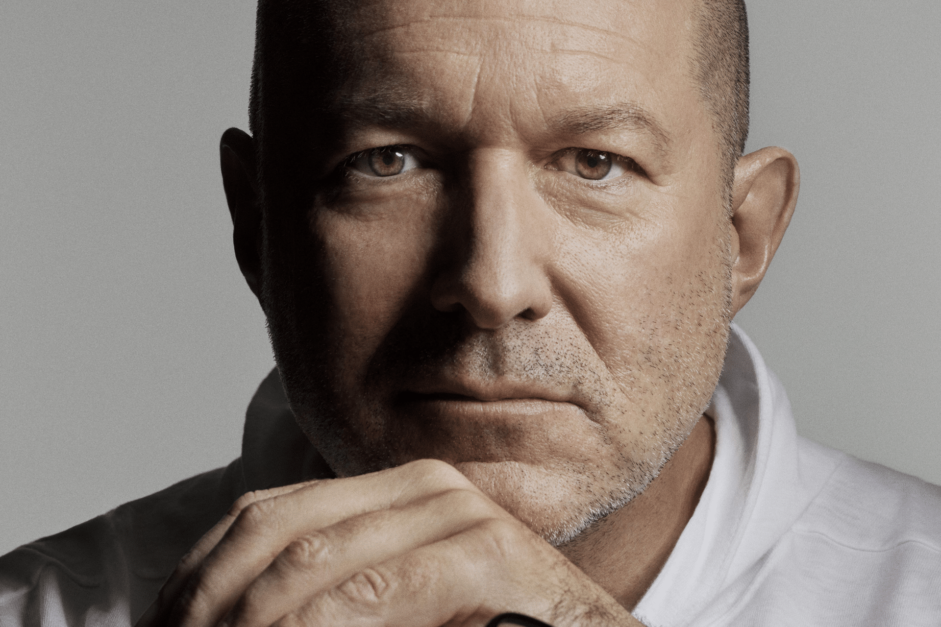 A middle-aged man with a bald head and stubble looks directly at the camera, resting his chin on his hands. Wearing a white shirt and a serious, contemplative expression, he evokes the thoughtful design seen in OpenAI's Jony Ive AI device concepts.