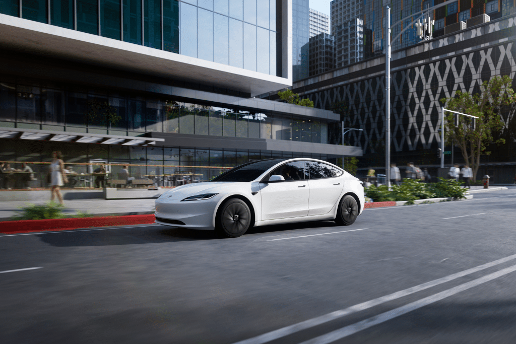 A sleek white 2026 Tesla Model 3 electric sedan glides down a city street lined with modern glass buildings, greenery, and outdoor seating on a sunny day.