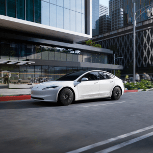 A sleek white 2026 Tesla Model 3 electric sedan glides down a city street lined with modern glass buildings, greenery, and outdoor seating on a sunny day.