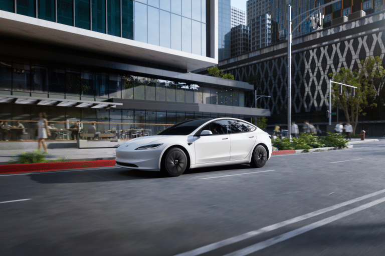 A sleek white 2026 Tesla Model 3 electric sedan glides down a city street lined with modern glass buildings, greenery, and outdoor seating on a sunny day.