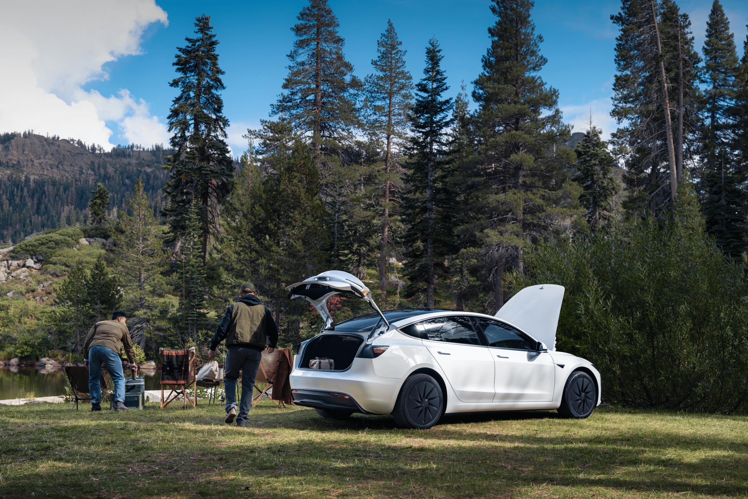A white 2026 Tesla Model 3 with open trunk and hood is parked on grass near a lake surrounded by tall pines; three people set up chairs by the water, enjoying a picnic in a scenic mountain landscape.
