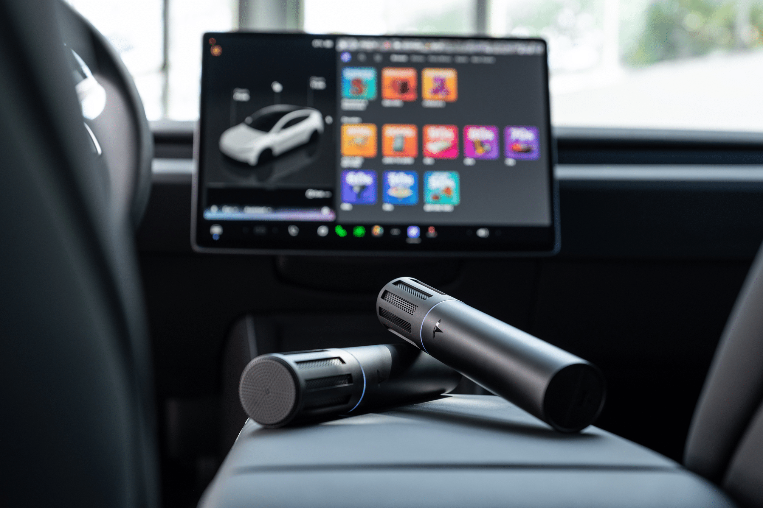 Two black, cylindrical wireless microphones rest on a 2026 Tesla Model 3 or Model Y Standard’s center console. In the background, a touchscreen display shows a car image and colorful app icons, hinting at in-car entertainment or karaoke features.
