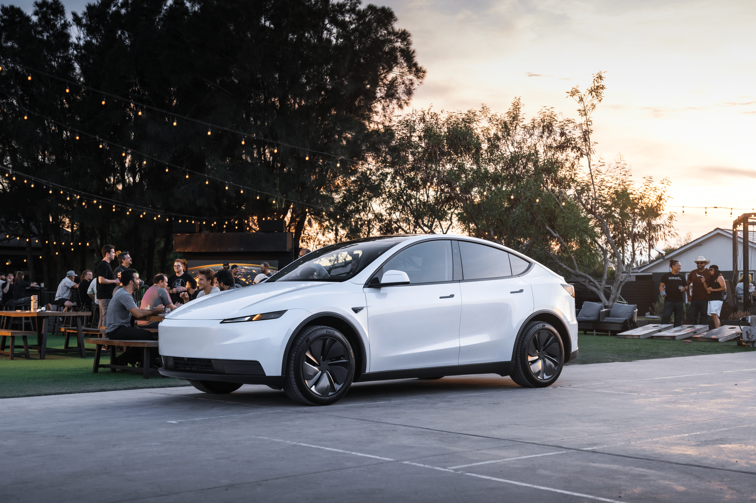 A white 2026 Tesla Model 3 and Model Y Standard parked outdoors near people sitting at picnic tables, with string lights and trees in the background during sunset.