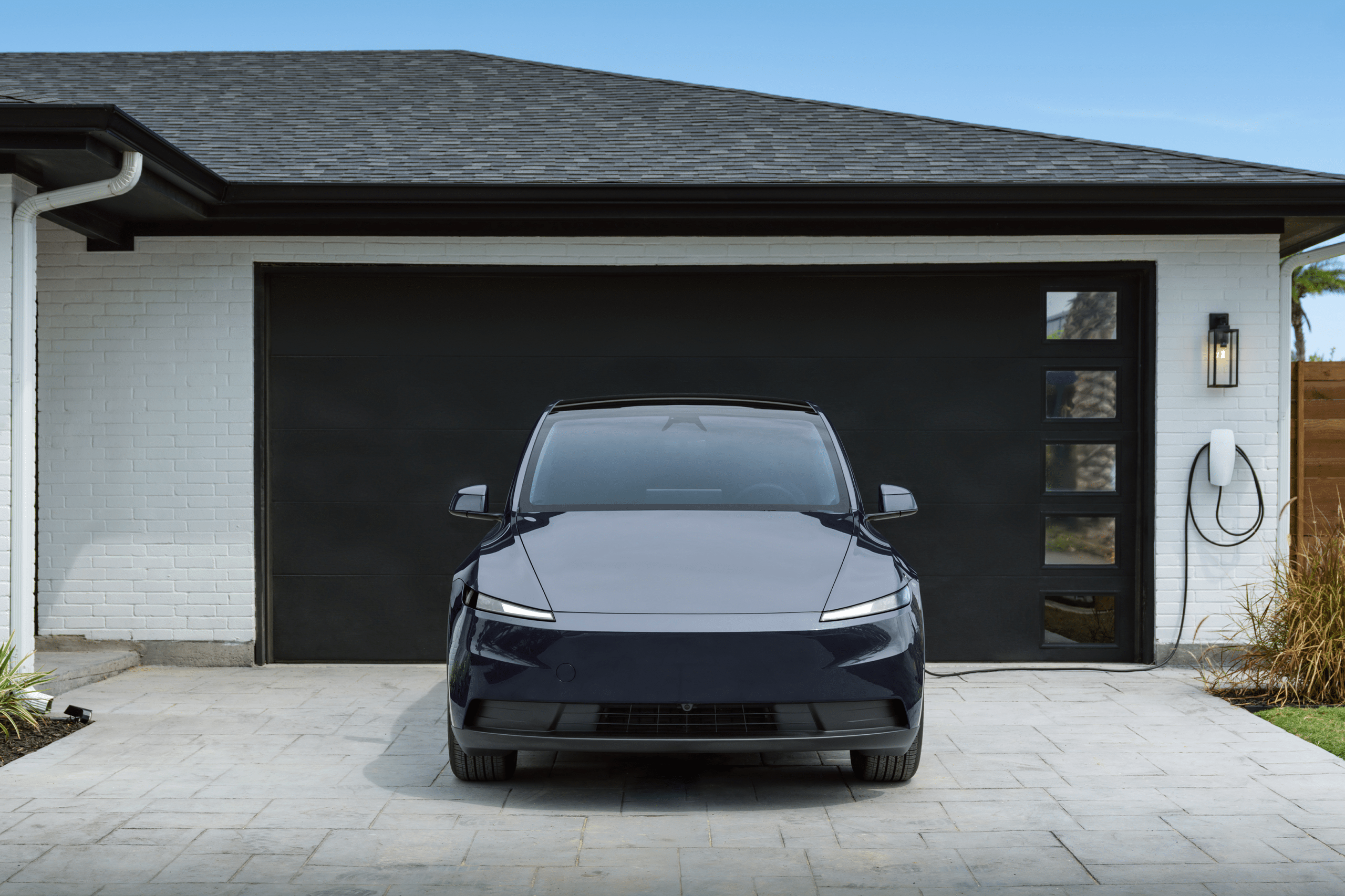 A sleek, dark 2026 Tesla Model 3 is parked in front of a modern house’s closed black garage door. An electric vehicle charging cable is mounted on the wall next to plants, with the light-colored tile driveway completing the scene.