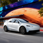 A sleek, silver 2026 Tesla Model 3 drives past a colorful mural featuring a large orange planet and abstract geometric shapes, with trees and buildings in the background.