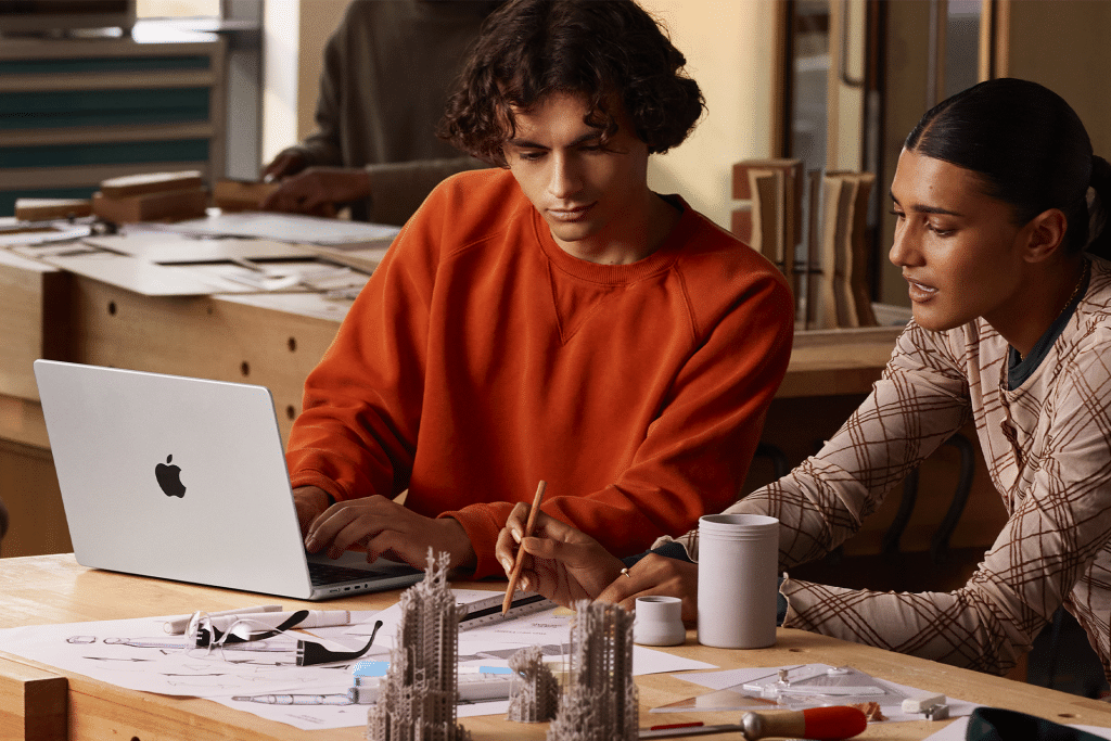 Two people sit at a table with architectural models and drawings, collaborating; one types on a 14-inch MacBook Pro M5 chip laptop while the other points at a sketch with a pencil.