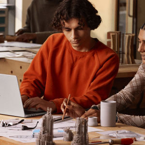 Two people sit at a table with architectural models and drawings, collaborating; one types on a 14-inch MacBook Pro M5 chip laptop while the other points at a sketch with a pencil.