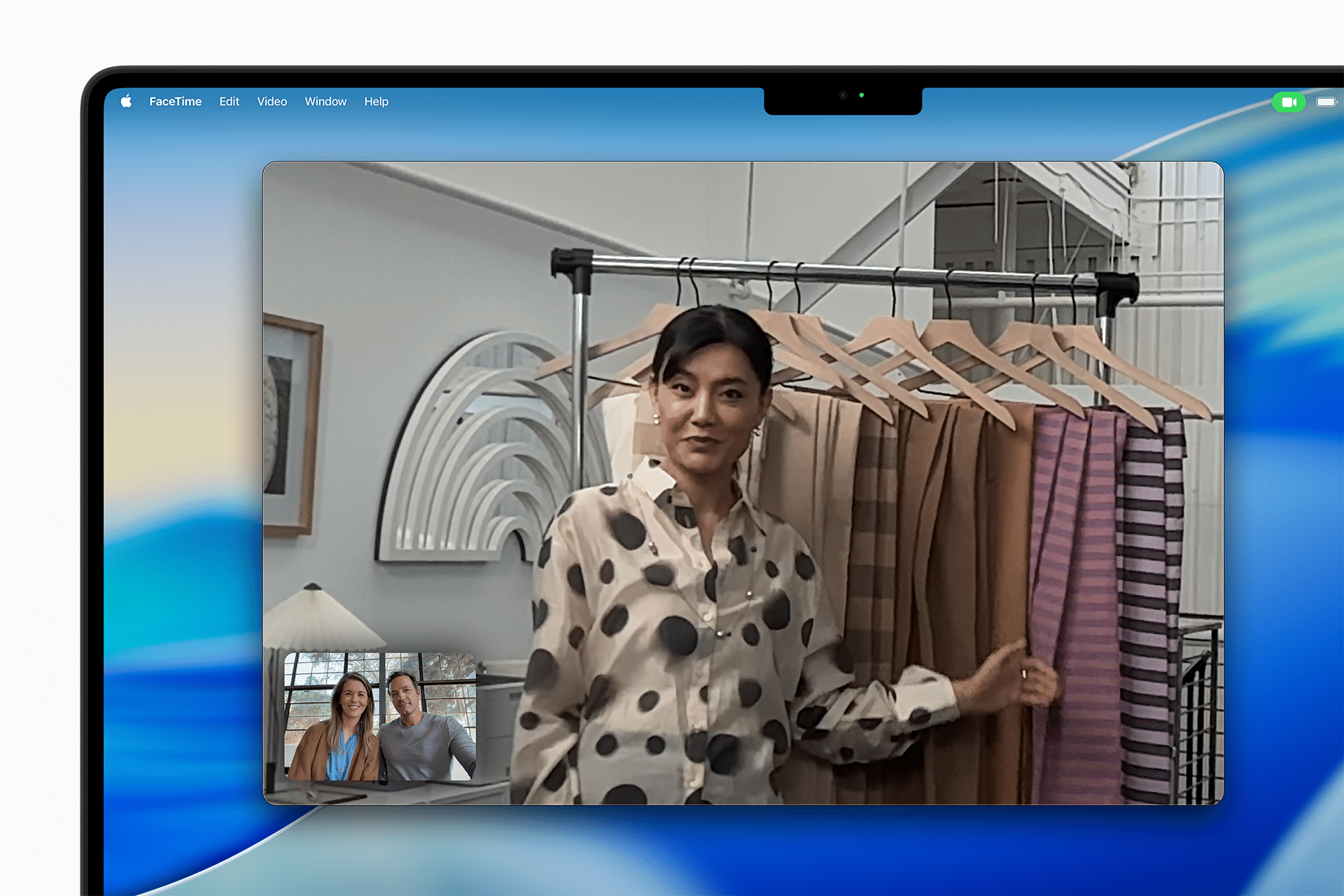 A woman stands by a clothes rack displaying various outfits during a video call on a MacBook Pro M5 vs M4. Two smaller video windows in the corner show a smiling couple. The room is bright and modern.
