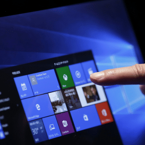 A hand is touching the screen of a tablet or computer displaying the Windows 10 start menu, highlighting colorful app tiles and shortcuts on a blue background, often discussed in debates about Microsoft unfair software pricing.
