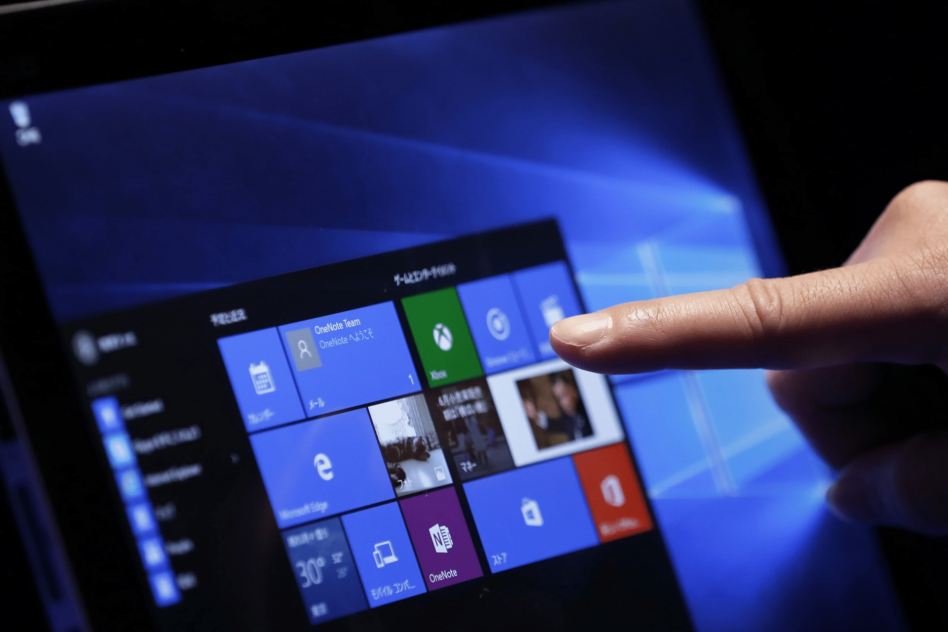 A hand is touching the screen of a tablet or computer displaying the Windows 10 start menu, highlighting colorful app tiles and shortcuts on a blue background, often discussed in debates about Microsoft unfair software pricing.