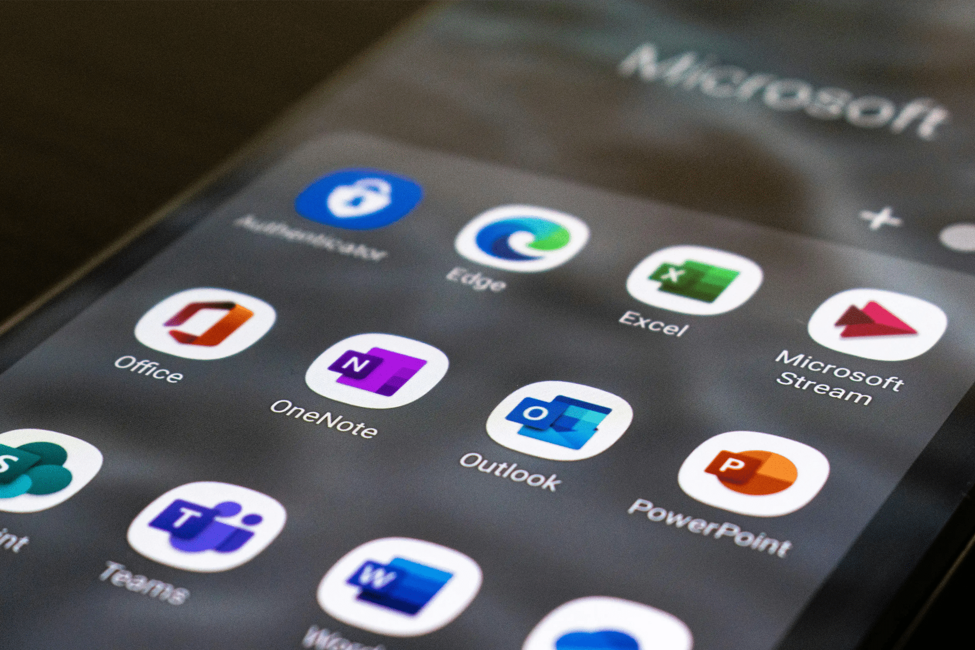 A smartphone screen displays Microsoft app icons, including Authenticator, Edge, Excel, Office, and Teams, arranged in a grid on a dark background—highlighting both the convenience and concerns over Microsoft unfair software pricing.