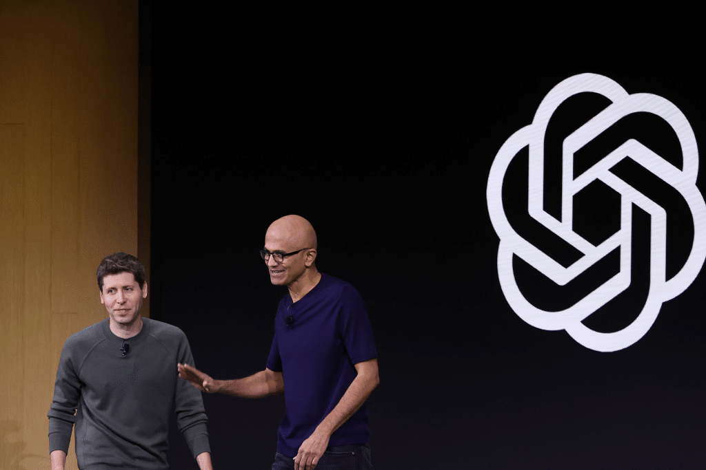 Two men stand on stage together, one in a green sweater and the other in a dark t-shirt, with a large OpenAI logo—fresh off headlines about its $500 billion valuation—displayed on a black background behind them.