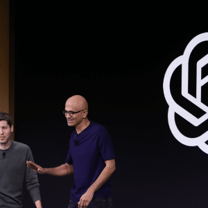 Two men stand on stage together, one in a green sweater and the other in a dark t-shirt, with a large OpenAI logo—fresh off headlines about its $500 billion valuation—displayed on a black background behind them.