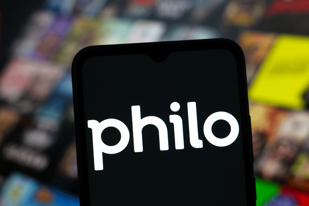 A smartphone screen displays the Philo logo in white text, with a blurred background featuring colorful, out-of-focus streaming content thumbnails, including references to Philo HBO Max favorites.