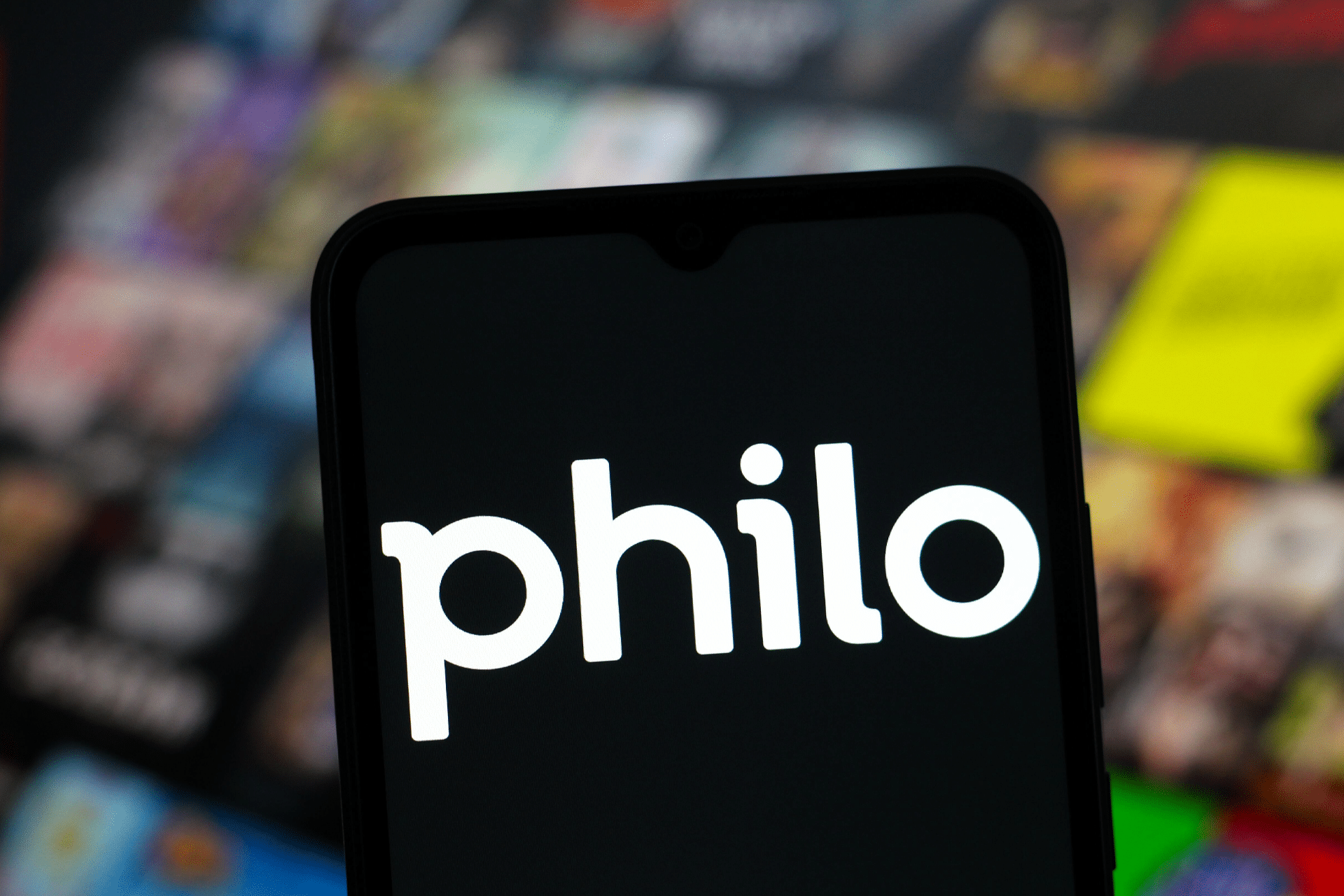 A smartphone screen displays the Philo logo in white text, with a blurred background featuring colorful, out-of-focus streaming content thumbnails, including references to Philo HBO Max favorites.