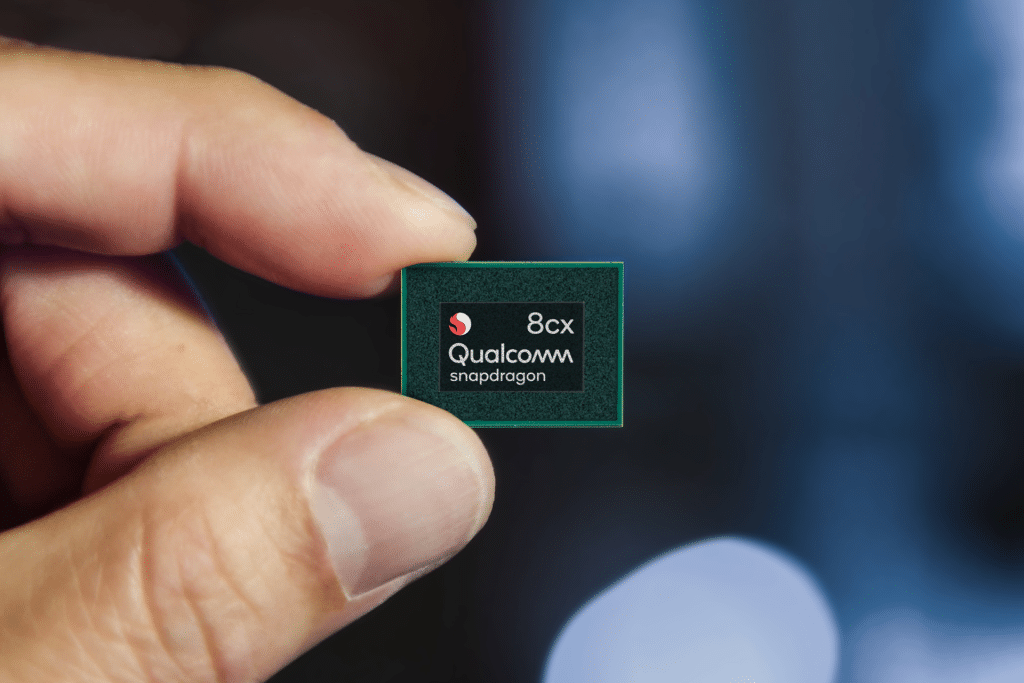 A person holds a small Qualcomm Snapdragon 8cx chip between their thumb and index finger, with a blurred blue and black background—symbolizing Qualcomm’s ongoing court battle over smartphone prices.