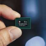 A person holds a small Qualcomm Snapdragon 8cx chip between their thumb and index finger, with a blurred blue and black background—symbolizing Qualcomm’s ongoing court battle over smartphone prices.